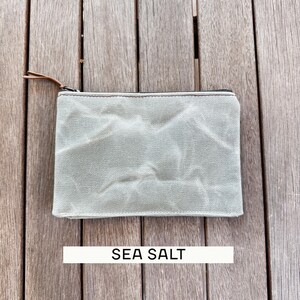 May include: A rectangular zippered pouch in a light sage green color with a textured, wrinkled fabric surface. The pouch features a dark metal zipper closure with a thin brown leather pull tab. The text SEA SALT is printed below the pouch.