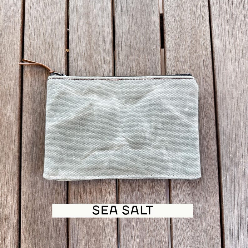May include: A rectangular zippered pouch in a light sage green color with a textured, wrinkled fabric surface. The pouch features a dark metal zipper closure with a thin brown leather pull tab. The text SEA SALT is printed below the pouch.