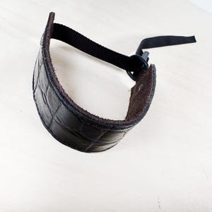 May include: A dark brown leather camera wrist strap with a crocodile skin texture. The strap has a black nylon webbing and a black plastic buckle for adjustment. The interior of the strap is a lighter brown color.