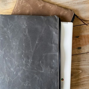 May include: A dark gray waxed canvas laptop sleeve with a black zipper closure, partially containing a white Lenovo Chromebook. A second brown waxed canvas zippered sleeve is positioned above the gray sleeve, both featuring a textured finish.