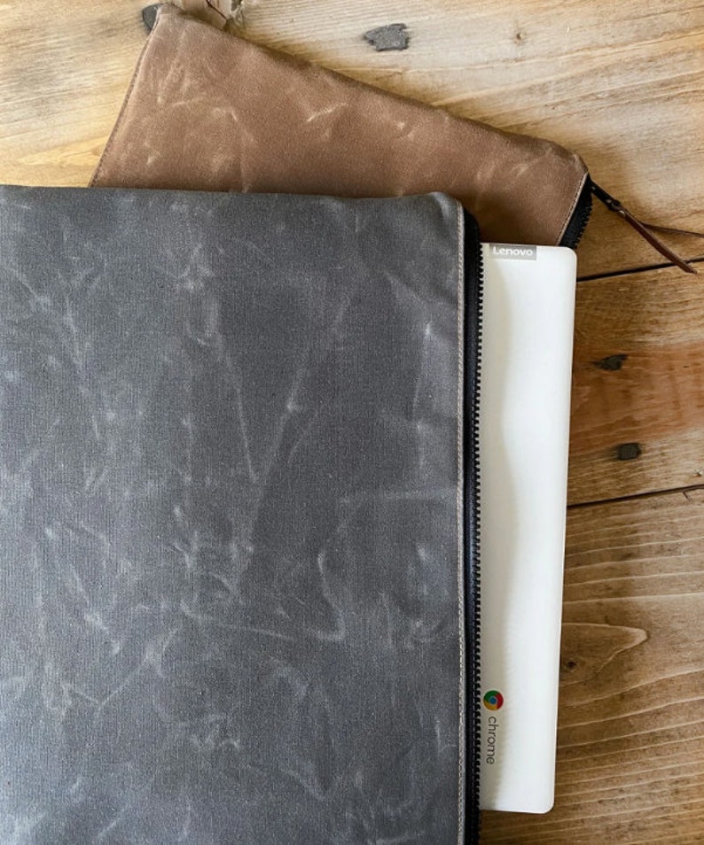 May include: A dark gray waxed canvas laptop sleeve with a black zipper closure, partially containing a white Lenovo Chromebook. A second brown waxed canvas zippered sleeve is positioned above the gray sleeve, both featuring a textured finish.