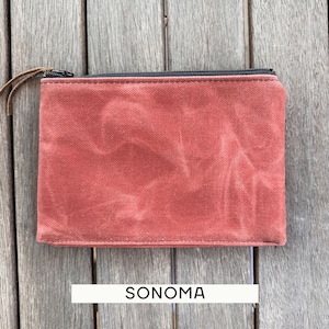 May include: A rectangular pouch made of a textured, rust-colored fabric with a black zipper closure along the top edge. A thin brown leather pull is attached to the zipper slider. The word SONOMA is printed in black text below the pouch.