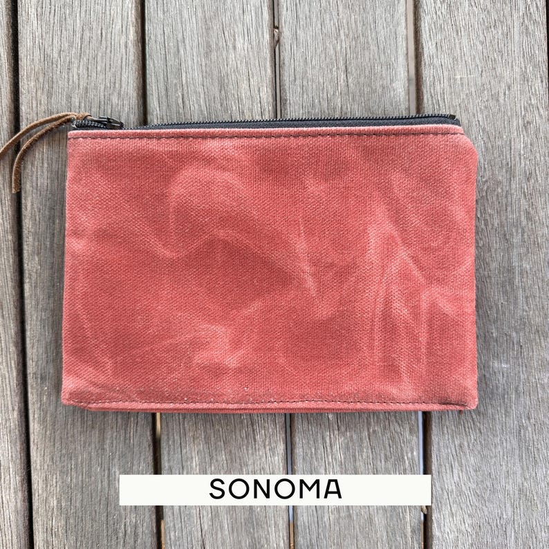 May include: A rectangular pouch made of a textured, rust-colored fabric with a black zipper closure along the top edge. A thin brown leather pull is attached to the zipper slider. The word SONOMA is printed in black text below the pouch.