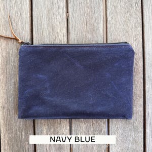 May include: A rectangular navy blue zippered pouch featuring a textured fabric surface and a brown leather pull tab attached to the metal zipper slider. The pouch has visible stitching along the top edge and the text NAVY BLUE printed below.