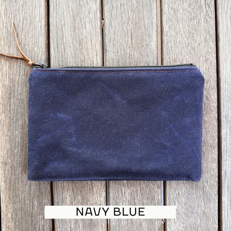 May include: A rectangular navy blue zippered pouch featuring a textured fabric surface and a brown leather pull tab attached to the metal zipper slider. The pouch has visible stitching along the top edge and the text NAVY BLUE printed below.