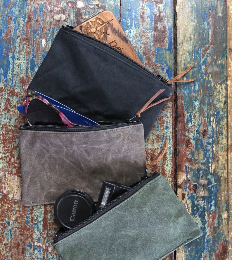 May include: Three rectangular zippered pouches in black, charcoal gray, and olive green. The black pouch contains a wooden item with a compass design. The gray pouch holds blue-framed sunglasses. The green pouch contains a black Canon lens cap.