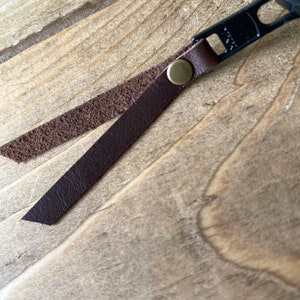 May include: A black metal zipper pull featuring a dark brown leather strap folded over and secured with a small brass-colored metal rivet. The leather strap has a smooth outer surface and a textured, rough inner side, showing the natural grain.