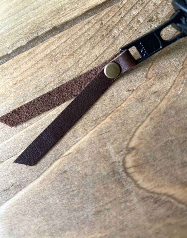 May include: A black metal zipper pull featuring a dark brown leather strap folded over and secured with a small brass-colored metal rivet. The leather strap has a smooth outer surface and a textured, rough inner side, showing the natural grain.