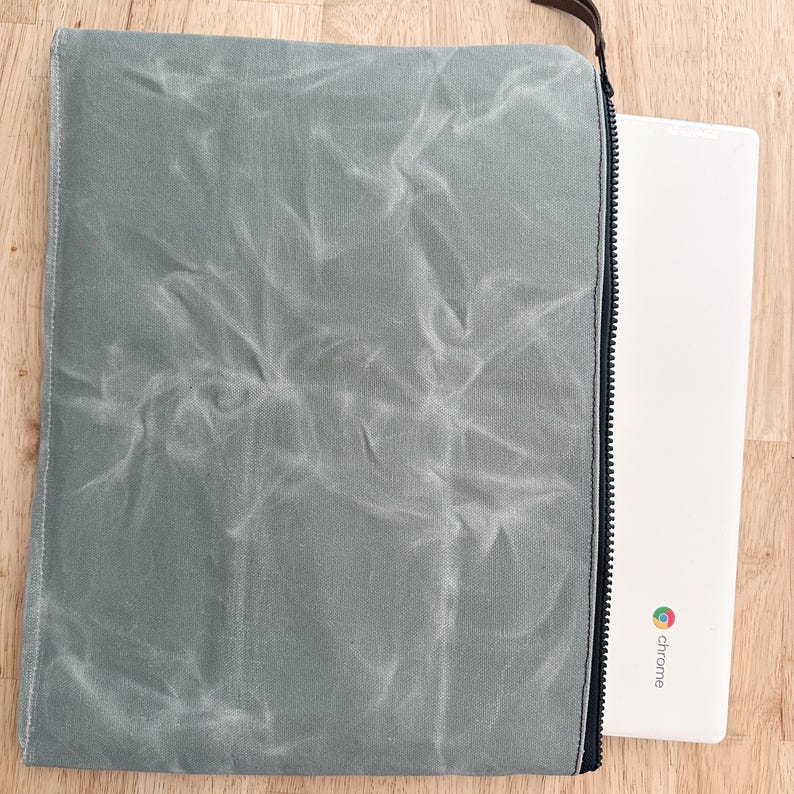 May include: A sage green waxed canvas laptop sleeve with a black zipper closure and a brown leather zipper pull. A white laptop with a colorful Chrome logo and the text chrome printed on the bottom corner is partially inserted into the sleeve.