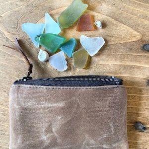 May include: A rectangular brown canvas zipper pouch with a dark brown leather pull tab. Above the pouch are several pieces of smooth, frosted sea glass in shades of light blue, green, olive, and amber, along with two small white spiral seashells.