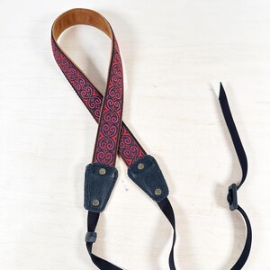 May include: A camera strap with a decorative pattern in red and dark purple. The strap has a brown leather backing and navy blue leather ends with brass rivets. Black nylon straps and a plastic clip are attached.