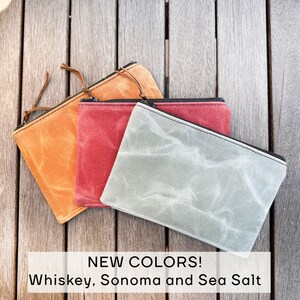 May include: Three rectangular zippered pouches in whiskey orange, Sonoma red, and sea salt gray. Each pouch features a textured, waxed canvas material with a black zipper closure and a thin leather pull tab. Text reads: NEW COLORS! Whiskey, Sonoma and Sea Salt.
