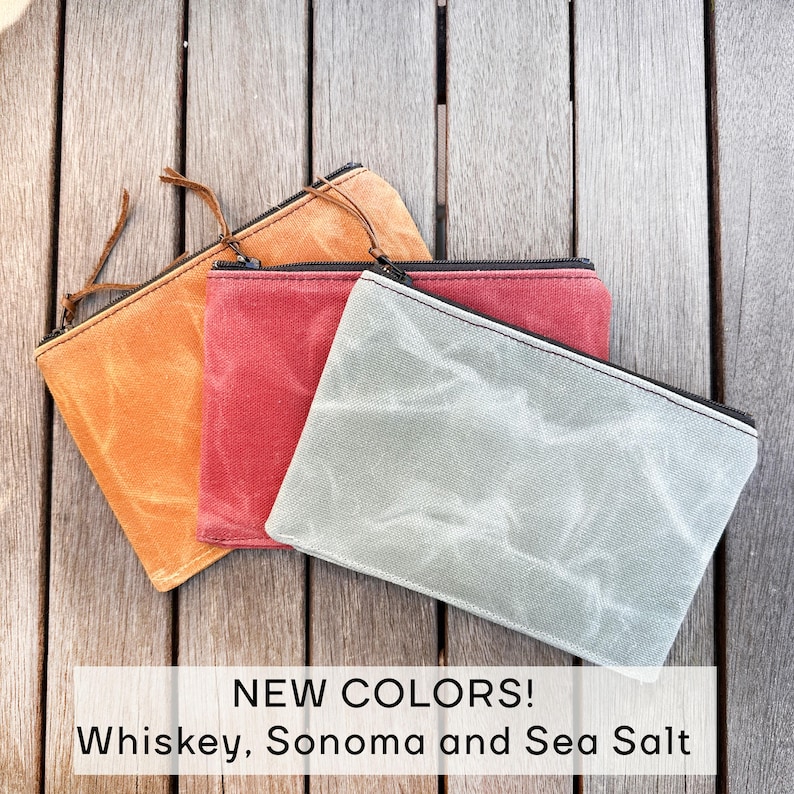 May include: Three rectangular zippered pouches in whiskey orange, Sonoma red, and sea salt gray. Each pouch features a textured, waxed canvas material with a black zipper closure and a thin leather pull tab. Text reads: NEW COLORS! Whiskey, Sonoma and Sea Salt.