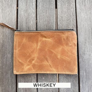 May include: A rectangular zippered pouch made of tan waxed canvas with a visible creased texture. The pouch features a black zipper closure along the top edge with a thin brown leather pull tab attached to the metal slider. The word WHISKEY is printed.