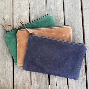 May include: Three rectangular zippered pouches in dark green, tan, and navy blue. Each pouch features a smooth, matte-textured fabric finish and a black zipper closure with a thin brown leather cord attached to the zipper pull for easy handling.