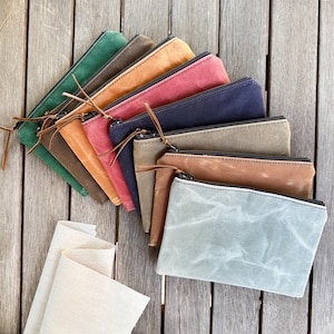 May include: A collection of rectangular zippered pouches arranged in a fan shape. The pouches are made of a textured, waxed fabric in various colors including green, brown, orange, red, navy blue, tan, light brown, and a light gray-blue.