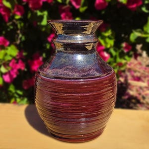 Könnte beinhalten: Handgefertigte Keramikvase mit glänzendem, dunkelburgunderfarbenem Finish. Die Vase hat ein strukturiertes, geripptes Design am unteren Teil und eine glatte, dunkle Metallic-Glasur am Hals. Es ist ein dekorativer Gegenstand.