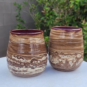 May include: Two handmade ceramic stemless wine glasses. The glasses have a brown and cream marbled pattern with a dark burgundy rim. The base of the glasses have a textured, off-white pattern. The glasses are displayed on a white surface.