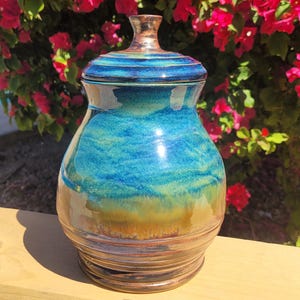 Pode incluir: Um pote de cerâmica artesanal com tampa. O pote apresenta uma vibrante mistura de tons turquesa, azul e castanho, com um acabamento brilhante. A tampa e a parte superior do pote têm listras horizontais. O pote está sobre uma superfície de madeira.
