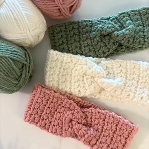 May include: Three handmade crochet headbands in sage green, cream, and dusty rose, with matching yarn balls in the background. The headbands have a textured stitch pattern and a twisted knot design.