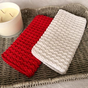 May include: A red and a white crocheted washcloth rest on a woven tray next to a white candle. The washcloths have a textured, raised pattern. The candle has three wicks and a light yellow wax.