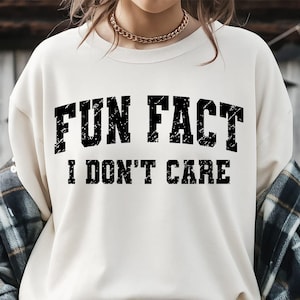 May include: Cream-colored sweatshirt with the text "FUN FACT I DON'T CARE" in distressed black lettering. The sweatshirt has a crew neck and long sleeves. A gold chain necklace is visible at the neckline.