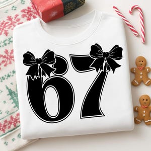 May include: White sweatshirt with the number 67 in black, each number adorned with a black bow. The sweatshirt is surrounded by holiday-themed decorations, including gingerbread men and candy canes.