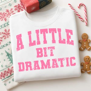 Könnte beinhalten: Weißes Sweatshirt mit dem Schriftzug „A LITTLE BIT DRAMATIC“ in rosa, verblichenen Buchstaben. Das Bild enthält festliche Dekorationen wie Lebkuchenmänner und Zuckerstangen, was auf ein Feiertags-Thema hindeutet.