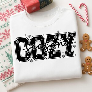 May include: White sweatshirt with the words "COZY SEASON" in a black, bold font with snowflake accents. The sweatshirt is folded, with a gift box, candy canes, and gingerbread cookies nearby. The overall theme is winter and the holidays.