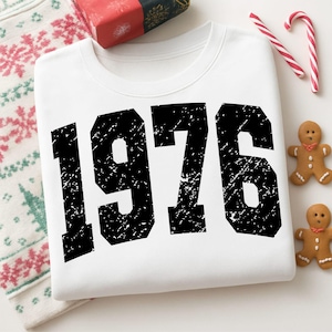 Può includere: Felpa bianca con l'anno "1976" stampato in grandi lettere nere effetto invecchiato. La felpa è piegata, con una confezione regalo, bastoncini di zucchero e omini di pan di zenzero nelle vicinanze. Il tema generale è vintage o retrò.