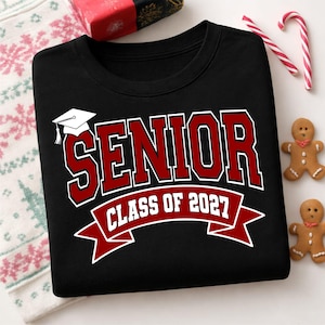 Könnte beinhalten: Ein schwarzes Sweatshirt mit Rundhalsausschnitt und einem Aufdruck, der das Wort SENIOR in großen roten Buchstaben mit weißer Umrandung zeigt, sowie ein Banner darunter mit der Aufschrift CLASS OF 2027. Ein weißes Doktorhut-Symbol ist über dem Wort SENIOR.