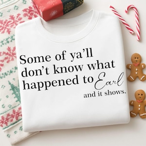 Pode incluir: Camisola branca com o texto "Some of ya'll don't know what happened to Earl and it shows." A camisola está dobrada com um padrão de tema natalino visível à esquerda. Dois bonecos de gengibre e bengalas de doces estão à direita.
