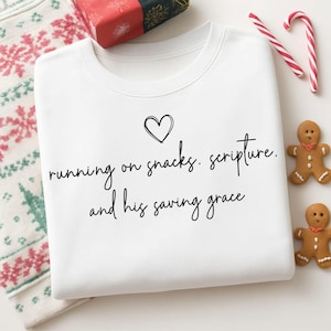 May include: White sweatshirt with the text "running on snacks. scripture. and his saving grace" in black script. A black heart is above the text. The sweatshirt is surrounded by Christmas-themed items, including candy canes and gingerbread men.