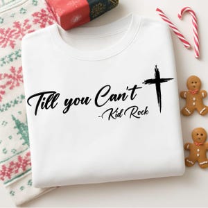 May include: White sweatshirt with the text "Till you Can't - Kid Rock" and a black cross graphic. The sweatshirt is folded and surrounded by holiday decorations, including candy canes and gingerbread cookies.