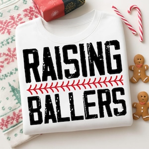 May include: White sweatshirt with the text "RAISING BALLERS" in black, with a red baseball stitch design. The sweatshirt is folded with a Christmas-themed pattern visible. Candy canes and gingerbread men are also present.