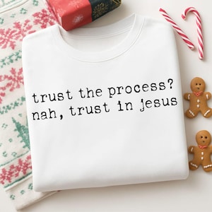May include: White crew neck sweatshirt with the text "trust the process? nah, trust in jesus" in black. The image includes a wrapped gift, gingerbread men, and candy canes.