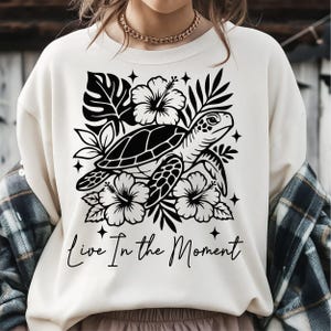 May include: A cream-coloured crewneck sweatshirt featuring a black graphic print of a sea turtle surrounded by hibiscus flowers, tropical leaves, and small four-pointed stars. The text "Live In the Moment" is printed in a cursive script below the graphic.
