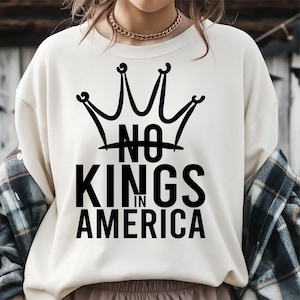 May include: Cream-colored sweatshirt with a black crown graphic and the text "NO KINGS IN AMERICA". The design is centered on the front of the shirt. The person is wearing a gold chain necklace and a plaid shirt.