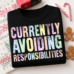 May include: A black crewneck sweatshirt featuring the text "CURRENTLY AVOIDING RESPONSIBILITIES" in a distressed, multi-colored pastel font. The letters are printed in shades of pink, yellow, blue, and green against the dark fabric background.