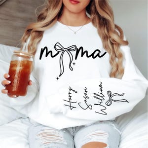 Op de afbeelding: Witte sweatshirt met het woord "mama" in zwart script, een strik-afbeelding en de namen "Harry", "Susan" en "William" op de mouw. Casual stijl.