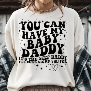 May include: Cream-colored sweatshirt with the text "YOU CAN HAVE MY BABY DADDY" in a black, retro font. Below, it reads "IT'S THE STEP DADDY I'LL CURB STOMP YOU FOR" with a heart graphic. The shirt has a casual style.