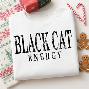May include: A white crewneck sweatshirt featuring the black text "BLACK CAT ENERGY" in a distressed, serif font. The garment is displayed alongside two gingerbread men cookies and two red and white striped candy canes on a patterned surface.