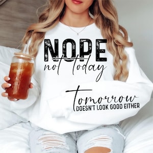 Könnte beinhalten: Weißes Sweatshirt mit dem Text "NOPE not today Tomorrow (doesn't look good either)" in Schwarz. Die Person trägt ein Sweatshirt und hält ein Glas Eistee. Das Sweatshirt hat einen lässigen Stil.