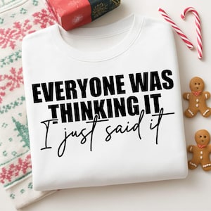 May include: White sweatshirt with the text "EVERYONE WAS THINKING IT, I just said it" in black. The image includes a wrapped gift, candy canes, and gingerbread cookies, suggesting a holiday theme.