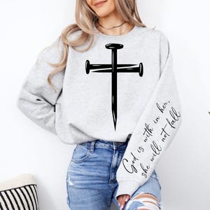Könnte beinhalten: Ein hellgraues Sweatshirt mit einem großen schwarzen Kreuzdesign aus Nägeln auf der Vorderseite. Der rechte Ärmel hat den Text "God is with in her, she will not fall" in schwarzer Schrift.