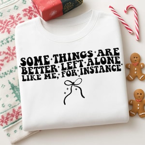 Puede incluir: Sudadera blanca con el texto negro "SOME THINGS ARE BETTER LEFT ALONE LIKE ME; FOR INSTANCE" y un lazo. También se ven decoraciones navideñas como bastones de caramelo y hombres de jengibre.
