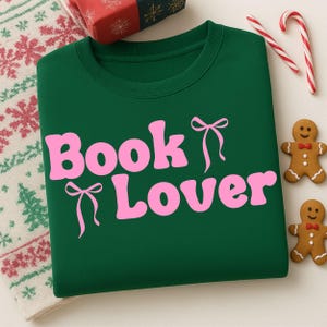 Może przedstawiać: Ciemnozielona bluza z napisem "Book Lover" w kolorze różowym, ozdobiona grafiką kokardek. Obraz zawiera pierniczki, laski cukrowe i zapakowany prezent, sugerując świąteczny motyw.