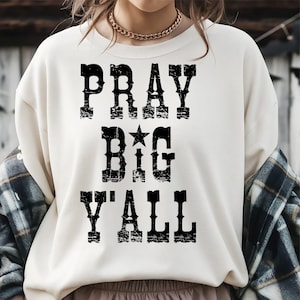 May include: Cream-colored sweatshirt with the text "PRAY BIG Y'ALL" in distressed black lettering. The word "BIG" features a star graphic. The sweatshirt is a casual, comfortable style.