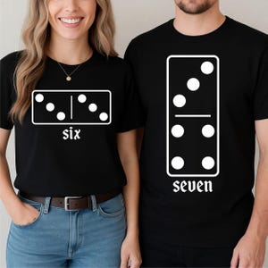 May include: Black t-shirts with white domino designs. One shirt displays a domino with six dots and the word "six." The other shirt shows a domino with seven dots and the word "seven."