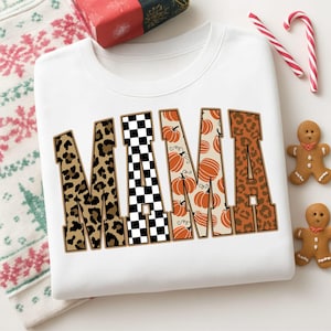 Könnte beinhalten: Ein weißes Sweatshirt mit Rundhalsausschnitt und dem Wort MAMA in großen Blockbuchstaben. Jeder Buchstabe hat ein eigenes Muster: Leopardenmuster, schwarz-weißes Schachbrettmuster, orangefarbene Kürbisse auf cremefarbenem Grund.